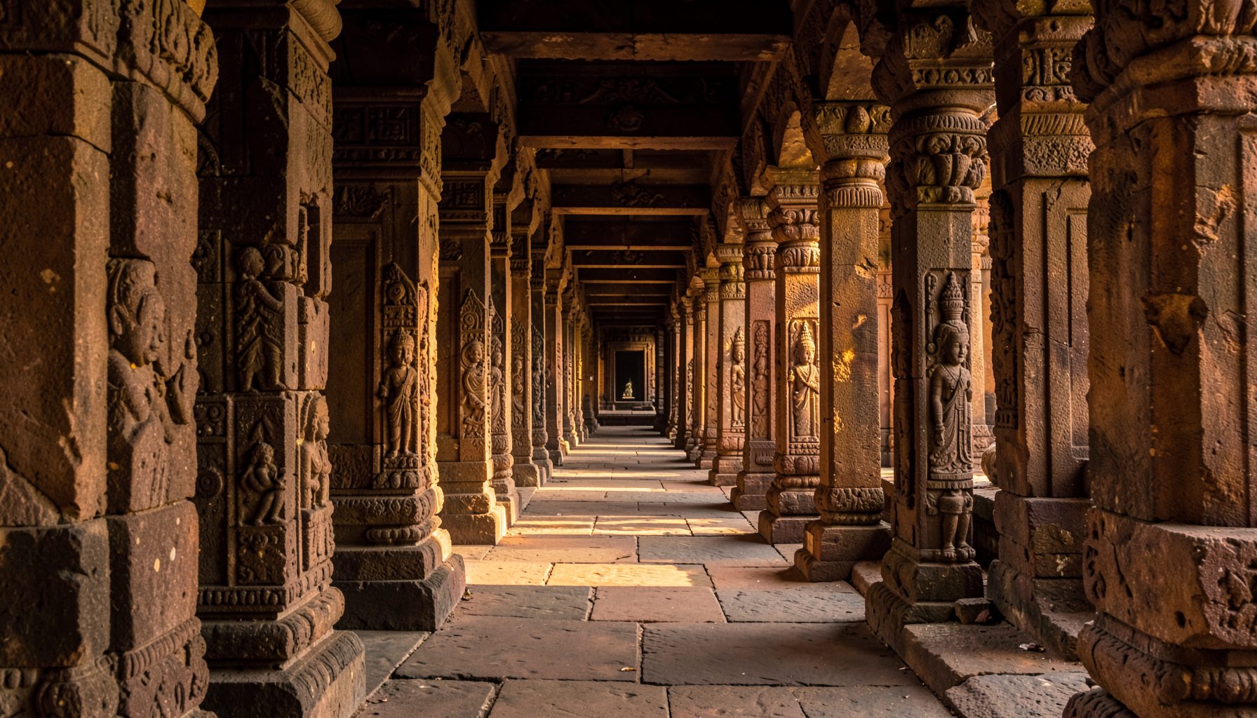 Ancient stone corridor of a South Indian temple with carved granite pillars and warm golden sunlight filtering through