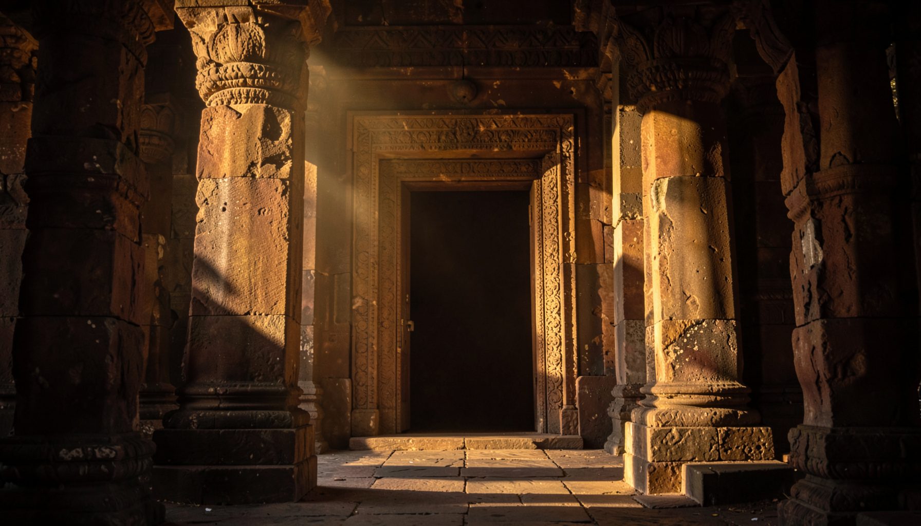Warm golden light falling across weathered stone pillars and a carved temple doorway, deep browns and ochres against a soft shadowed interior